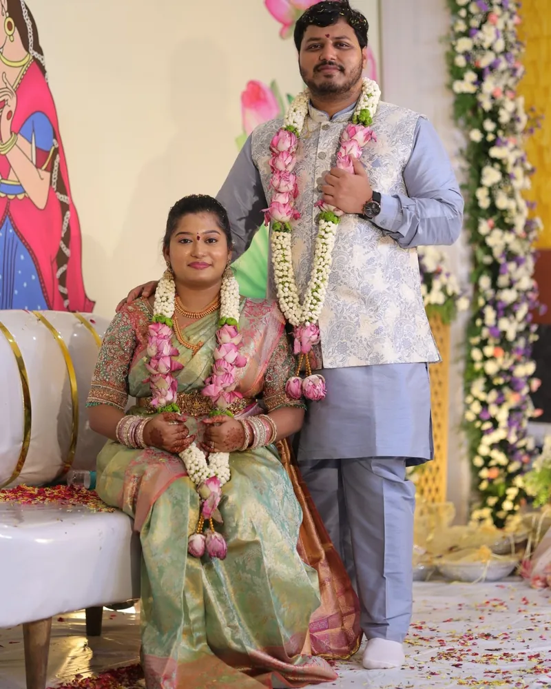 Susmita seated and Ashish standing beside her, both wearing garlands